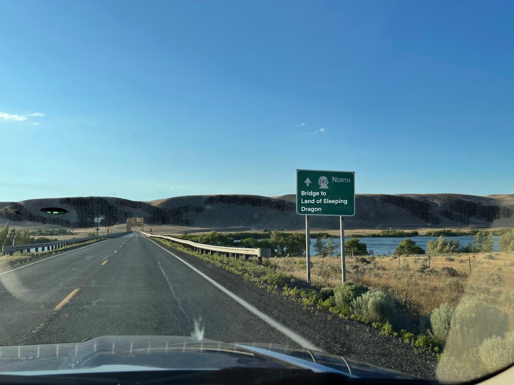 Highway sign before bridge over river to rolling hills with a half open eye on the left hill: Bridge to Land of Sleeping Dragon
