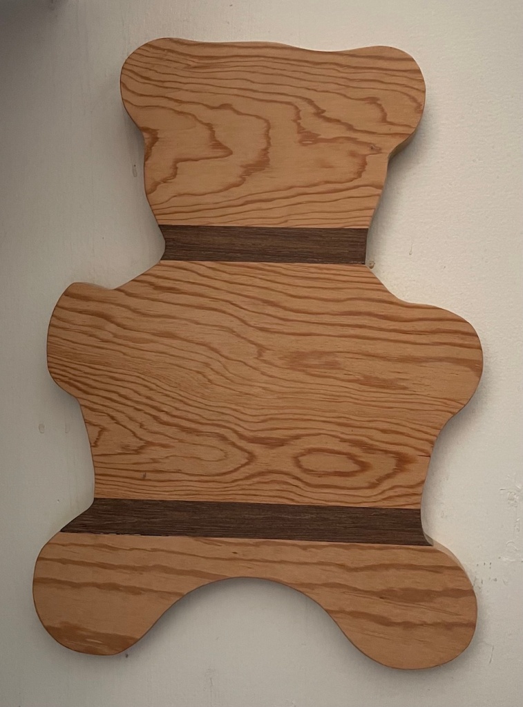 hanging on a white wall is a handcrafted wooden cutting board in the shape of a bear with two darker wooden strips at neck and waist